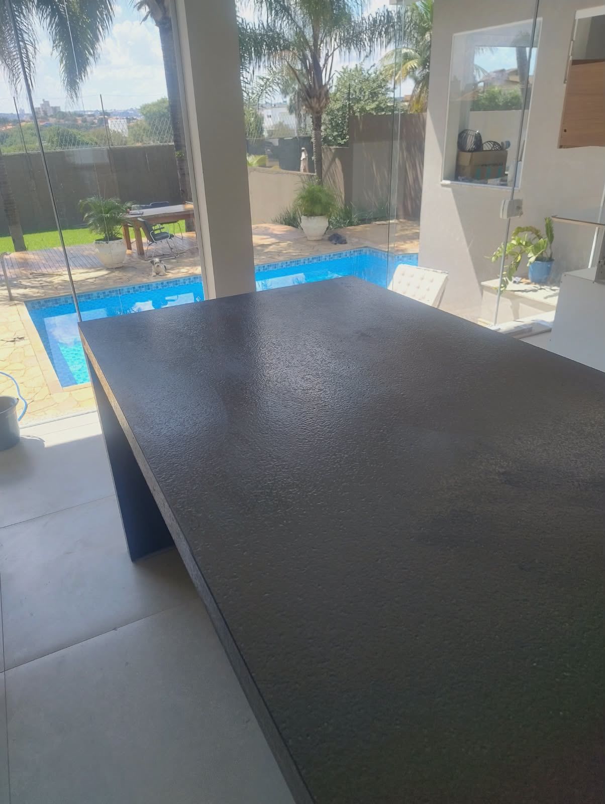 Ilha de granito escuro com acabamento texturizado, piscina e jardim ao fundo
