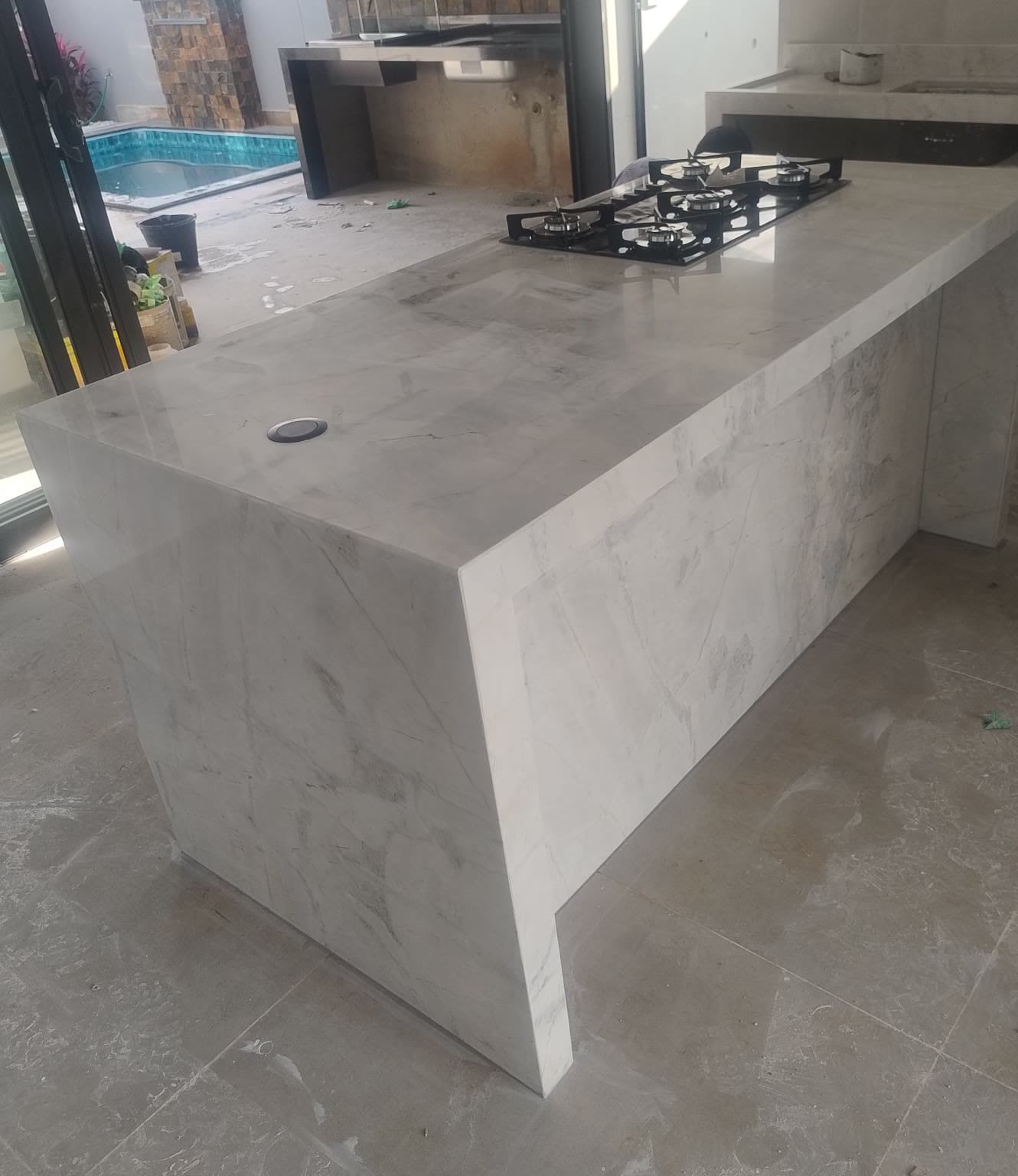 Ilha de pedra clara com cascata e cooktop, com a piscina ao fundo